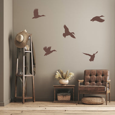 Flying duck wall decals in brown vinyl displayed on a neutral-toned wall above a rustic wooden chair and side table in a cozy cabin-style room.